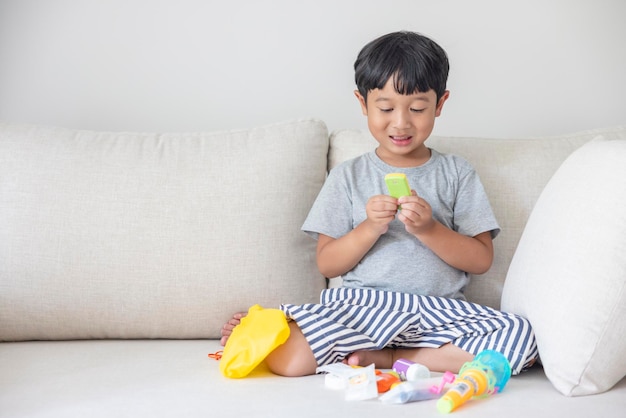 Entzückender glücklicher asiatischer kleiner Junge, der ein graues Hemd und blau-weiß gestreifte Shorts trägt, die auf einem cremefarbenen Sofa sitzen, lächelt glücklich und wählt seine Spielsachen absichtlich aus