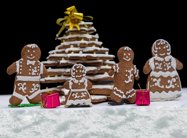 Entzückender hausgemachter Lebkuchen-Weihnachtsbaum mit süßer Lebkuchenfamilie mit Geschenken auf dunklem Hintergrund. Mockup für saisonale Angebote und Urlaubspostkarte