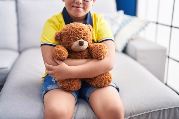 Entzückender hispanischer Junge, der den Teddybär umarmt, der zu Hause auf dem Sofa sitzt