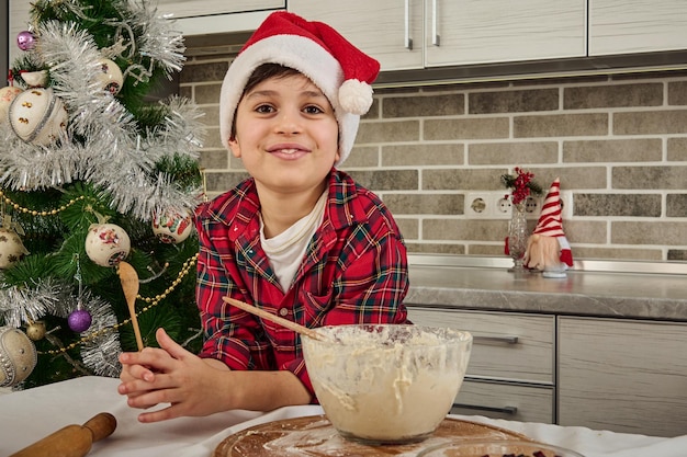 Entzückender hübscher vorpubertärer Junge in kariertem Hemd und Weihnachtsmütze hält einen Holzlöffel und steht am Küchentisch mit einer Schüssel mit Weihnachtsplätzchenteig, lächelt zähnefletschend in die Kamera