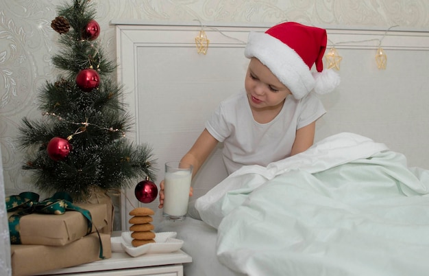 Entzückender Junge kocht Milch und Kekse für den Weihnachtsmann, Freiraum. Ein traditioneller Leckerbissen ist ein Weihnachtskeks und ein Glas Milch auf dem Nachttisch neben dem Bett. Das Konzept des neuen Jahres. Verwandte