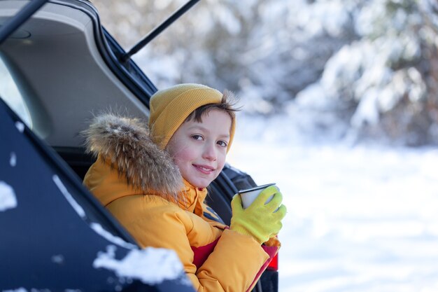 Entzückender Junge mit heißem Tee oder Kakao in den Händen, der am Wintertag im schwarzen Auto sitzt. Roadtrip, Kurzurlaub, natürliche Umgebung, Aufenthalt, Reisen, Tourismus im Winter.