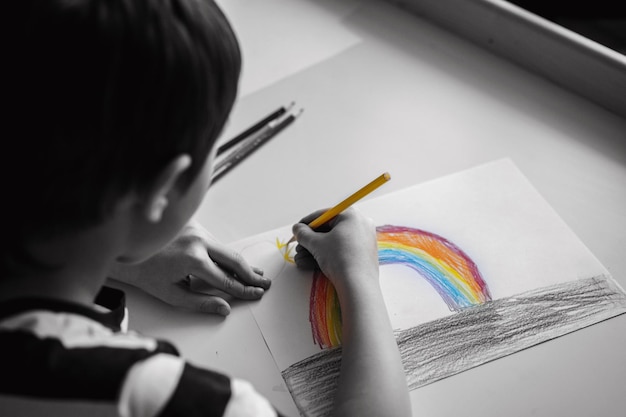 Entzückender kaukasischer Junge im Grundschulalter, der einen Regenbogen mit Bleistiften zeichnet. Schwarz und weiß