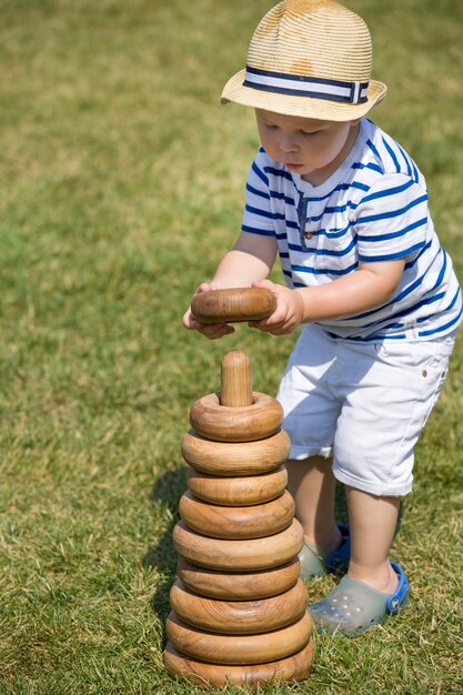 Foto entzückender kleiner junge, der an einem sonnigen tag mit einer großen hölzernen spielzeugpyramide auf dem gras im freien spielt