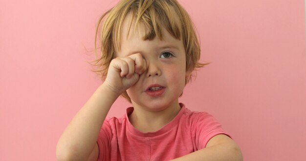 Entzückender kleiner Junge im rosa T-Shirt, welches das Auge schaut schläfrig reibt