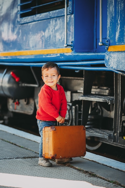 Entzückender kleiner Junge in rotem Pullover auf einem Bahnhof in der Nähe des Zuges mit Retro-altem braunem Koffer. Bereit für den Urlaub. Junge Reisende auf dem Bahnsteig.