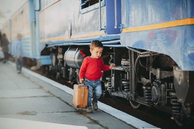 Entzückender kleiner Junge in rotem Pullover auf einem Bahnhof in der Nähe des Zuges mit Retro-altem braunem Koffer. Bereit für den Urlaub. Junge Reisende auf dem Bahnsteig.