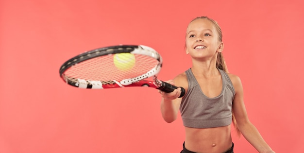 Entzückender Mädchen-Tennisspieler, der ein Sport-Crop-Top trägt, während er Tennisball auf dem Schläger hüpft und lächelt