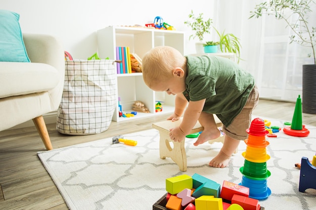 Entzückender und aktiver blonder Junge, der seine Körperfunktionen erforscht und versucht, richtig auf einer kleinen Holzbank zu sitzen Kinder trainieren ihre neuen Fähigkeiten Kinderzimmereinrichtung mit weißen Möbeln und vielen Spielsachen