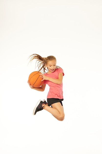 Entzückender weiblicher Kinderbasketballspieler, der Spielball hält und schreit, während er in die Luft springt. Isoliert auf weißem Hintergrund