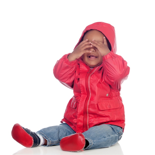 Entzückendes afrikanisches Baby, das auf dem Boden mit dem roten Regenmantel bedeckt das Gesicht sitzt