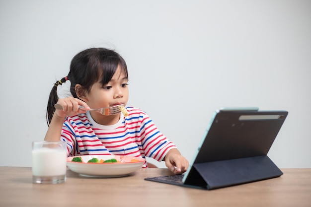 Entzückendes asiatisches Mädchen, das zu Mittag isst, während es sich einen Film von der Tablette ansieht Ein kleines asiatisches Kind, das zu Abend isst, und die Augen sehen aus der Tablet National Eating Disorders Awareness Week