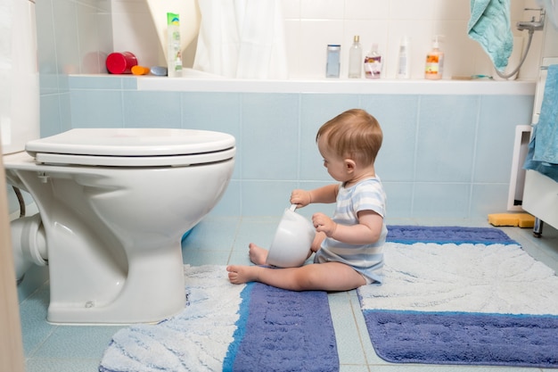 Entzückendes Baby, das im Badezimmer auf dem Boden sitzt und mit Toilettenpapier spielt