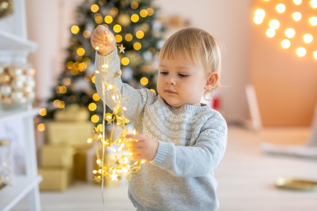 Entzückendes Baby, das in der Nähe des Weihnachtsbaums mit festlichen Lichtern und Weihnachtsgeschenken Weihnachten und Neujahr sitzt