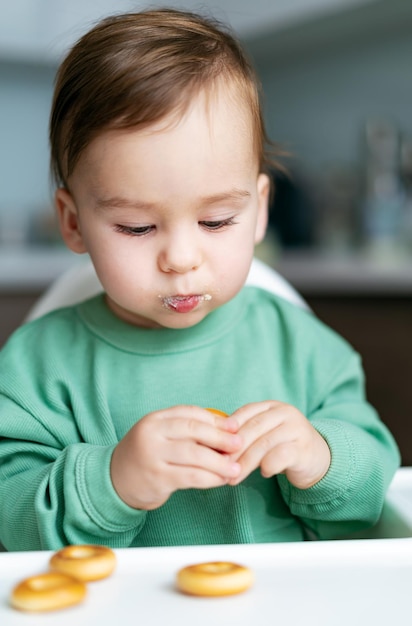 Entzückendes Baby, das Plätzchen isst, die im Hochstuhl sitzen Baby, das Nahrung auf Küche isst