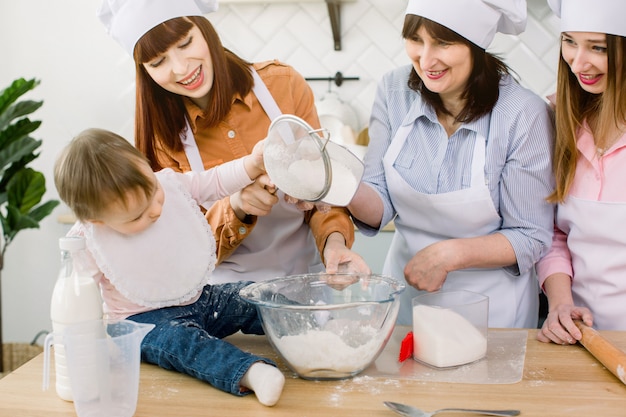 Entzückendes Baby mit Mutter, Tante und Großmutter, die Teig zu Hause mit Mehl und Ei und Zucker zusammen machen. Frauen in weißen Schürzen und Kochmützen streuen Teig für Gebäck mit Mehl auf Tisch