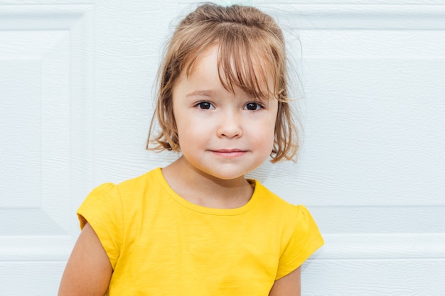 Entzückendes blondes Mädchen, das ein gelbes Hemd trägt, das gegen weißen Hintergrund lehnt