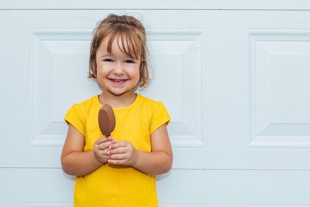 Entzückendes blondes Mädchen, das Eis isst und ein gelbes Hemd trägt, das sich gegen weißen Hintergrund lehnt