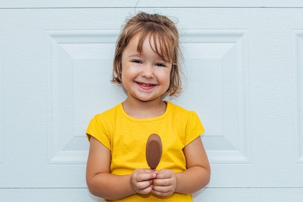 Entzückendes blondes Mädchen, das Eis isst und ein gelbes Hemd trägt, das sich gegen weißen Hintergrund lehnt