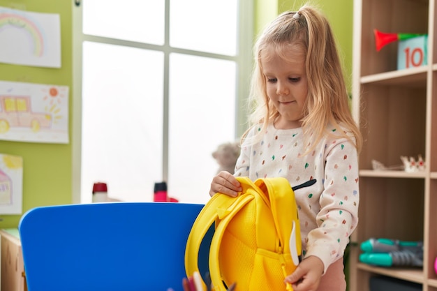 Foto entzückendes blondes mädchen im vorschulalter lächelt selbstbewusst beim öffnen des rucksacks im kindergarten