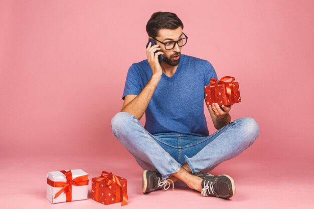 Entzückendes Foto des attraktiven jungen Mannes mit dem schönen Lächeln, das Geburtstagsgeschenkkästen lokalisiert über rosa Wand hält, die auf dem Boden sitzt. Geschenkkonzept. Telefon benutzen.