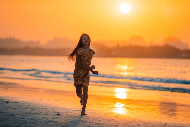 Entzückendes glückliches kleines Mädchen am weißen Strand bei Sonnenuntergang.