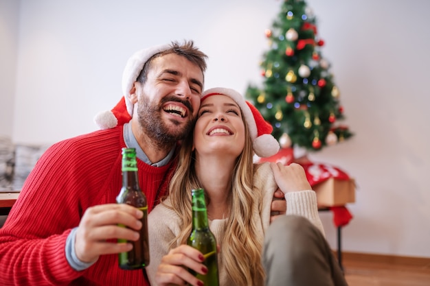 Entzückendes gutaussehendes kaukasisches Paar mit Weihnachtsmützen auf Köpfen, die auf Boden im Wohnzimmer sitzen, Bier umarmen und halten. Im Hintergrund ist Weihnachtsbaum mit Geschenken.
