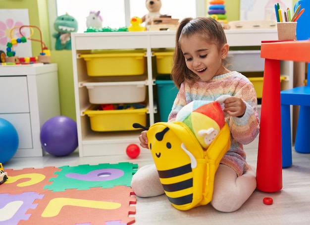 Entzückendes hispanisches Mädchen im Vorschulalter, das im Kindergarten auf dem Boden sitzt und einen Ball aus Rucksack hält