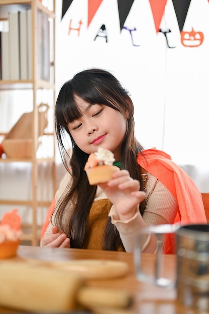 Entzückendes junges asiatisches Mädchen im Halloween-Kostüm, das einen Halloween-Kürbiskleinen Kuchen hält