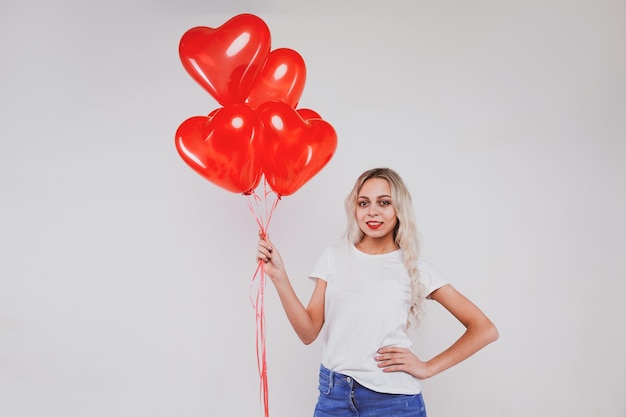 Entzückendes junges blondes Mädchen in Jeans und weißem T-Shirt posiert mit herzförmigen Luftballons auf weißem Hintergrund Valentinstag-Konzept