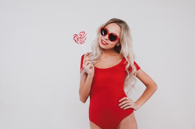Entzückendes junges blondes Mädchen in rotem Body und Brille in Form von Herzen, die auf weißem Hintergrund posieren
