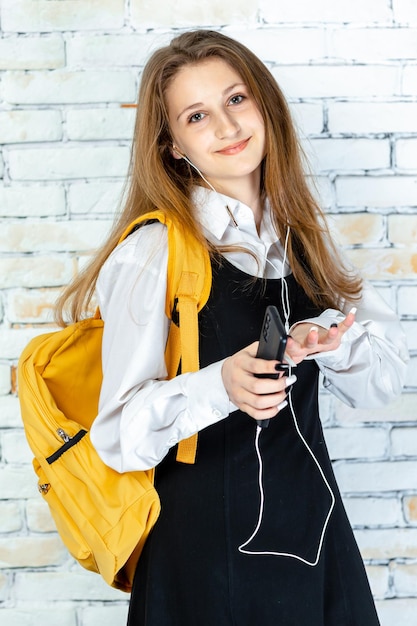 Entzückendes junges Mädchen in Schuluniform, das ihr Telefon hält und Kopfhörer verwendet Foto in hoher Qualität