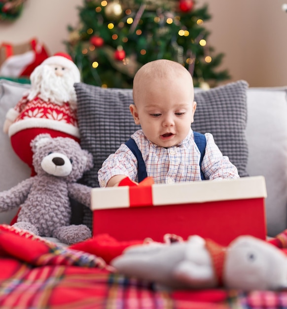 Entzückendes kaukasisches Baby, das ein Geschenk hält, das zu Hause auf dem Sofa am Weihnachtsbaum sitzt