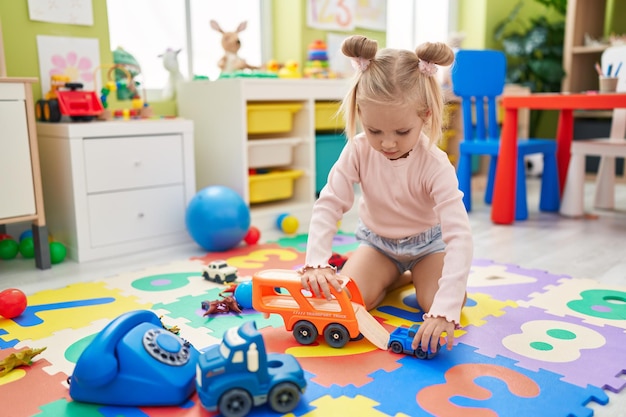 Entzückendes kaukasisches Mädchen, das mit Autospielzeug spielt, das im Kindergarten auf dem Boden sitzt