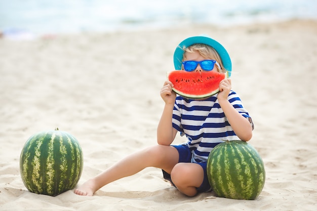 Entzückendes Kind am Seeufer, das saftige Wassermelone isst. Fröhliches Kind im Sommer am Strand. Netter kleiner Junge draußen