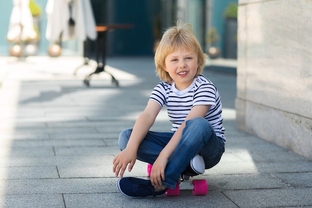 Entzückendes Kind im Freien. Nettes hübsches Kind, das in die Kamera lächelt. Zufälliger Junge auf der Sommerzeit, die auf einem Skateboard skatet.