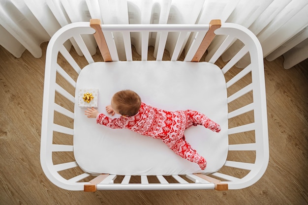 Entzückendes Kind im Weihnachtspyjama mit Geschenken, die zu Hause im Kinderbett liegen. Baby mit Weihnachtsgeschenken in der Wiege im Babyzimmer