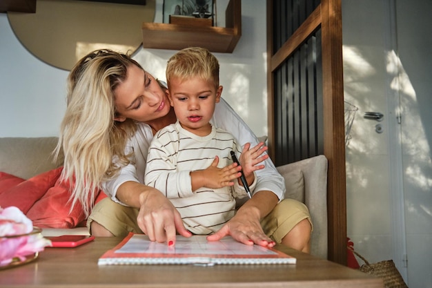 Entzückendes Kind mit Stift, das interessiert auf das Lehrbuch schaut und lesen lernt, während es neben der liebevollen Mutter steht, die zu Hause auf dem Sofa im Wohnzimmer sitzt