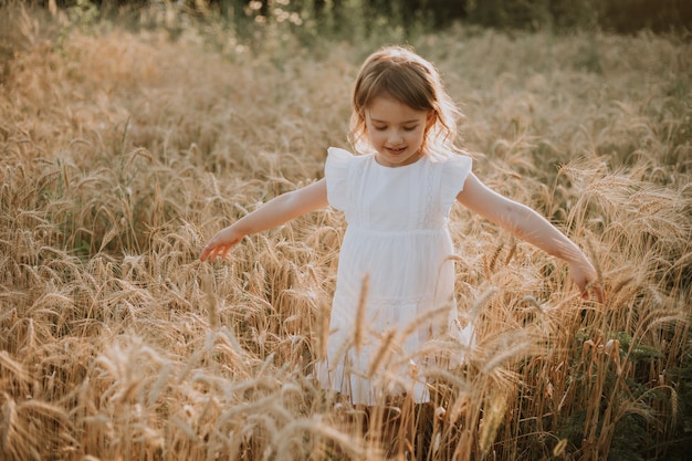 Entzückendes kleines blondes Mädchen in einem Hut und einem weißen Leinenkleid, das auf einem Weizenfeld steht