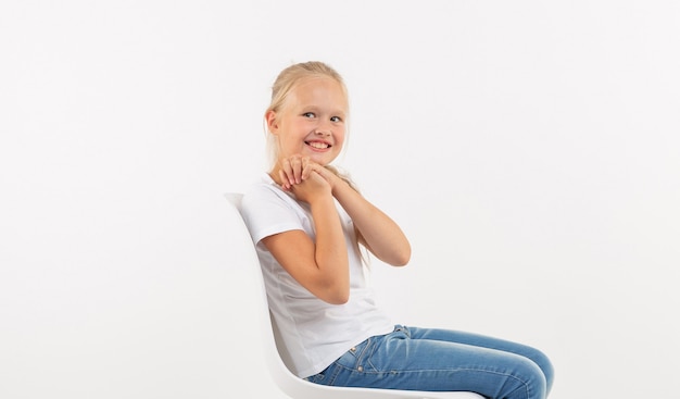 Entzückendes kleines Kindermädchen mit dem langen blonden Haar, das auf dem Stuhl auf weißem Hintergrund sitzt.