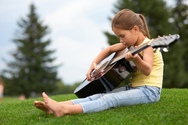 Entzückendes kleines Mädchen, das auf einem grünen Gras im Park sitzt und Gitarre hält und Zeit mit den Eltern im Freien verbringt