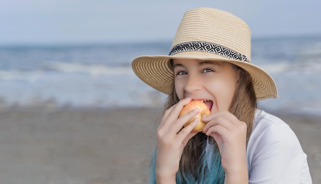 Entzückendes kleines Mädchen, das einen Apfel in einem Cowboyhut am Strand isst