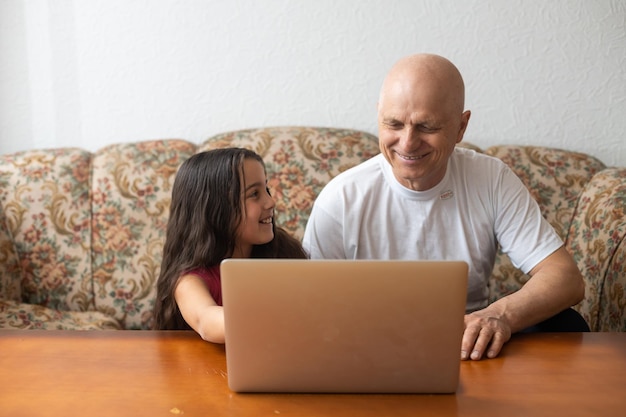 Entzückendes kleines Mädchen, das glücklichen Großvater mit Laptop zu Hause umarmt.