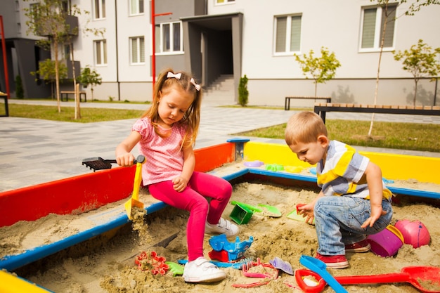 Foto entzückendes kleines mädchen, das in einem sandkasten spielt, junge, der auf die spielsachen schaut