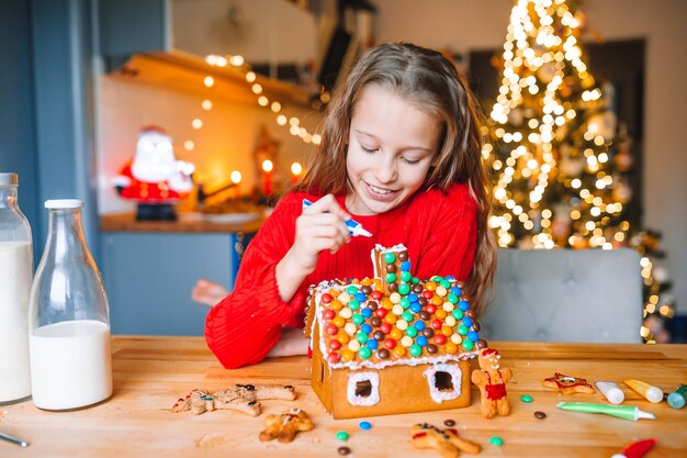 Entzückendes kleines mädchen, das lebkuchenhaus mit glasur verziert. schöne küche mit lichtern und weihnachtsbaum, tisch mit kerzen. frohe weihnachten und schöne feiertage