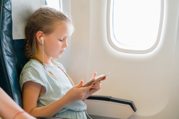 Entzückendes kleines Mädchen, das mit einem Flugzeug reist. Nettes Kind mit Laptop nahe Fenster in den Flugzeugen