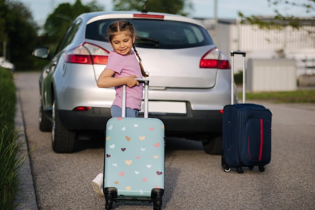 Entzückendes kleines Mädchen, das mit seinem Kinderkoffer vor dem Auto steht und auf die Familienreise der Eltern wartet