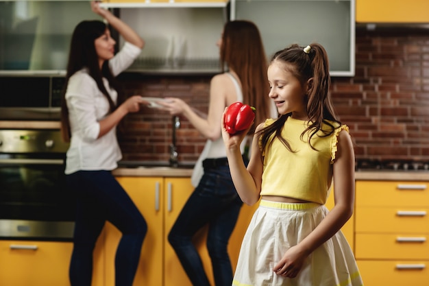 Entzückendes kleines Mädchen, das Paprika hält und es betrachtet. Glückliches Mädchen, das in der Küche steht und mit einem großen Paprika spielt. Im Hintergrund stapeln zwei Frauen Geschirr. Werbung für gesunde Ernährung