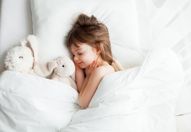 Entzückendes kleines Mädchen, das süß in einem weißen Bett mit Kaninchenspielzeug schläft, das nahe liegt. Draufsicht.