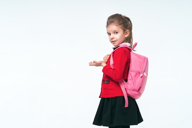 Entzückendes kleines Mädchen in der roten Schuljacke, im schwarzen Kleid, an den Trägern eines Rucksacks und lächelnd und in die Kamera schauend, auf weißem Raum posierend. Isolieren